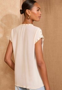 Blusa color crema con maniche corte, caratterizzata da dettagli in oro sulle spalle, realizzata in tessuto leggero e morbido.