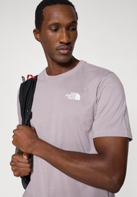 Ανοιχτό μωβ κοντομάνικη μπλούζα με κοντό λαιμόκοψη, που φέρει λευκό λογότυπο στο στήθος. Άτομο κρατάει το λουράκι μιας μαύρης τσάντας πλάτης.