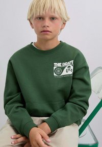 Sudadera de algodón verde con un diseño gráfico de baloncesto y el texto "THE DRAFT". Puños acanalados, cuello redondo y detalle de bolsillo canguro.