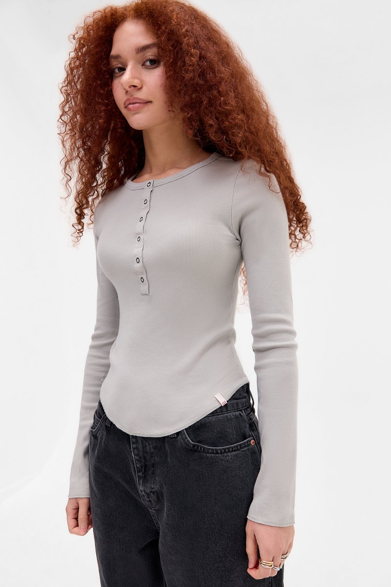 Lys grå ribbet langermet topp med knappestolpe og buet kant, parret med mørk grå jeans og en liten merkeetikett på siden.