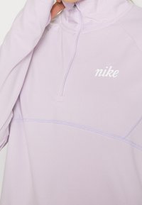 Pullover Nike di colore lilla chiaro con colletto alto, chiusura a mezza zip e dettagli cuciti. Logo bianco sull'area del petto sinistro. Tessuto morbido e liscio.