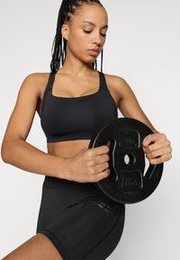 Zwarte atletische sportbeha met een vierkante halslijn en brede bandjes, gecombineerd met zwarte shorts met hoge taille, die een zwart gewichtsschijf vasthoudt.