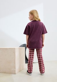 T-shirt ample à manches courtes de couleur violette associée à un pantalon évasé à carreaux rouge et beige ; présenté sur un fond minimaliste.