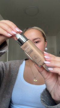 Bottiglia di fondotinta L'Oréal True Match con corpo in vetro trasparente, pompa argentata, liquido beige e etichette stampate, tenuta da una persona.