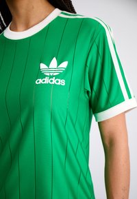 Jersey verde de Adidas con franjas verticales negras, cuello blanco y tres franjas blancas en los hombros. Logo blanco prominente en el pecho.