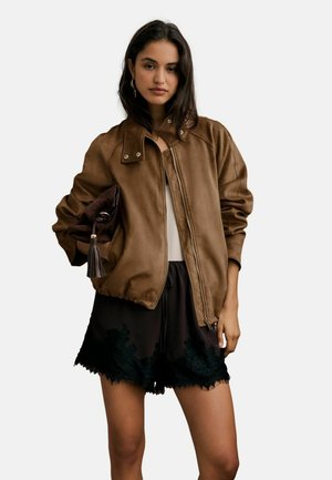 Frau mit langen dunklen Haaren, die eine braune Reißverschlussjacke und schwarze Shorts mit Spitzenbesatz trägt, hält eine braune Clutch mit Quaste unter dem Arm.