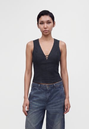 Mujer con cabello corto y negro lleva una camiseta negra sin mangas con escote en V profundo y vaqueros azules de pierna ancha, de pie frente a un fondo gris liso.