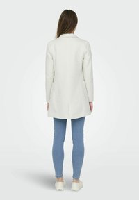 Witte wolblendjas met een getailleerde pasvorm, enkelvoudig knoopsluiting, en twee zijzakken, gecombineerd met lichtblauwe skinny jeans en witte schoenen.