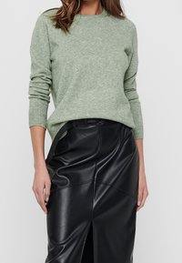 Hellgrüner Strickpullover mit gerippten Bündchen und Saum, kombiniert mit einem schwarzen Kunstlederrock mit einem Schlitz vorne und glatter Textur.