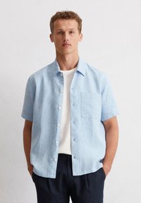 Helles blaues Kurzarmhemd aus Leinen mit durchgehender Knopfleiste und einer Brusttasche, getragen über einem weißen T-Shirt.