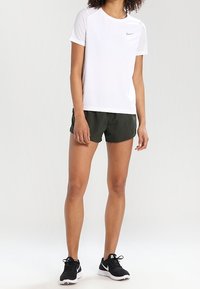 Mujer vestida con una camiseta blanca de manga corta de Nike, pantalones cortos verdes oscuros y zapatillas de correr negras de Nike de pie contra un fondo neutro.