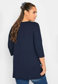 Camicia tunica blu navy con maniche a tre quarti, texture liscia e spacchi laterali per facilità di movimento. Indossata con jeans di denim.