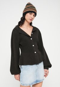 Σκούρα καφέ ριγωτή ζακέτα με φαρδιά γιακά και πέντε λευκά κουμπιά, σε συνδυασμό με φούστα από ανοιχτό μπλε τζιν. Το μοντέλο φοράει πολύχρωμο σκουφάκι.