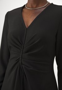 Abito nero con maniche lunghe, scollatura a V e dettaglio arricciato sul davanti. Tessuto liscio con una leggera lucentezza, indossato con una collana di perle argentate.