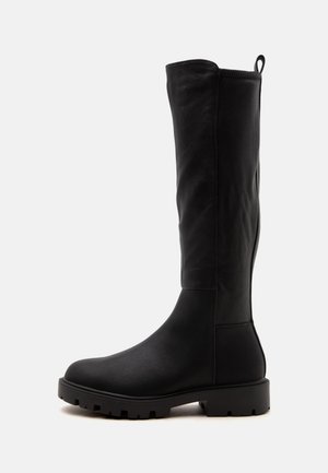 Botte noire montante jusqu'au genou avec semelle à gros crampons et languette à l'arrière, vue de profil sur un fond clair uni.