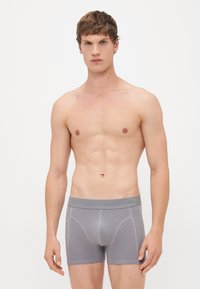 Fiatal férfi áll egy fehér háttér előtt, illeszkedő szürke boxer alsóneműt viselve, amelynek derékrészén a "JACK & JONES" felirat látható, előrefelé nézve semleges kifejezéssel az arcán.