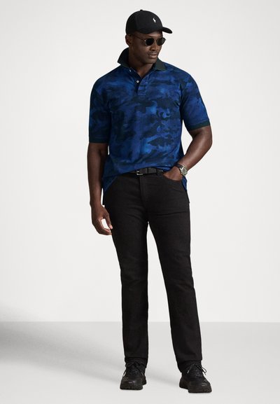 Mężczyzna w niebieskiej koszulce polo w kamuflażu, czarnych dżinsach, czarnych trampkach, czarnej czapce, okularach przeciwsłonecznych i zegarku na nadgarstku stojący z jedną ręką w kieszeni.