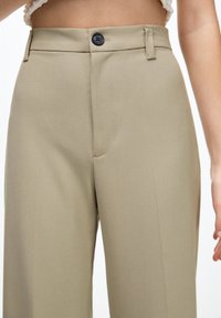 Pantaloni alti beige chiaro con piega frontale, chiusura con un bottone e due tasche laterali. Tessuto liscio e strutturato.