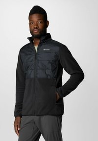 Giacca in pile nera con un pannello frontale trapuntato nero. Presenta un collo alto e chiusura a zip. Texture liscia, design leggero.