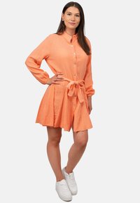 Abito button-up in tessuto peach texturizzato, con maniche lunghe, vita legata e gonna svasata. Abbinato a sneakers bianche.