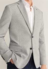 Lichtgrijs colbert van gestructureerde stof. Heeft een enkele knoop, revers met inkeping en zijzakken. Draagt over een wit overhemd.