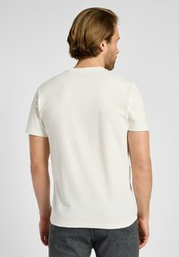 Camiseta de manga corta en algodón color blanco roto, con cuello redondo y un acabado suave y texturizado. La vista trasera muestra un branding mínimo y un diseño sencillo.
