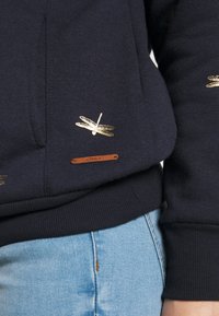 Felpa blu navy con ricamo di libellula dorata e un'etichetta in pelle marrone con la scritta "ONLY". Presenta un design strutturato e polsini a costine.