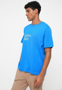 Mėlyna trumpomis rankovėmis marškinėlių su apvalia iškirpte ir priekyje esančiu geltonu "calvin klein" logotipu. Pagaminti iš minkštos, lengvos medžiagos, laisvai krentantys.