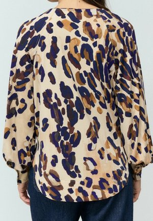 Blouse beige avec imprimé léopard abstrait bleu, marron et beige, manches longues bouffantes et poignets boutonnés, portée avec un jean foncé.