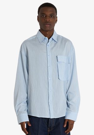 Uomo che indossa una camicia azzurro chiaro a righe sottili con maniche lunghe e una grande tasca frontale, in piedi davanti a uno sfondo bianco uniforme.