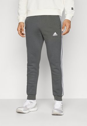 Grå joggingbukser med hvide sidelstriber og et adidas-logo på venstre hofte. Fremstillet af blødt materiale, med en tætsiddende pasform og manchetter ved anklerne.