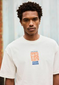 Fiatal férfi göndör hajjal, fehér pólót visel, amelyen narancssárga és kék grafikával "Loquat and Bird Utagawa Hiroshige" felirat található a mellkasán.