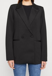 Mustassa kaksoisnappisella bleiserillä, jossa on hammastetut kaulukset, ryhdikkäät olkapäät ja kaksi etutaskua. Sileää kangasta; minimalistikotulostus.