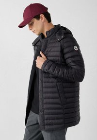 Giacca a vento nera imbottita con trapuntatura orizzontale, cappuccio e tasche laterali. Indossata sopra una maglietta nera, abbinata a un cappello bordeaux.