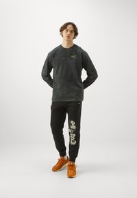 Hosszú ujjú, sötétszürke Nike póló zöld logóval, fekete jogger nadrággal, amely fehér grafikával díszített. Narancssárga sportcipő egészíti ki az öltözetet.