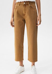 Personne portant un jean marron court avec des ourlets bruts et des baskets blanches, debout devant un fond blanc.