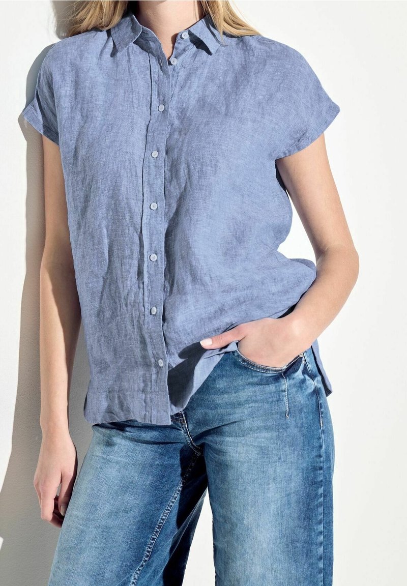 Femme portant une chemise bleu clair à manches courtes boutonnée et un jean bleu, une main dans la poche, sur fond blanc.