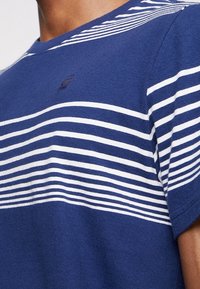 Marinblå bomullströja med horisontella vita ränder och en liten logotyp på bröstet. Klassisk modell med rund halsringning och korta ärmar.