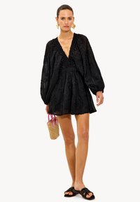 Vestido negro bordado con un escote en V profundo, mangas texturizadas y una silueta fluida, combinado con sandalias planas y un bolso de mimbre.