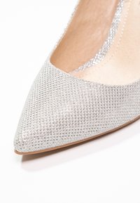 Sølv højhælet sko med spidse tæer, dækket af små glitrende studs, med en glat inderside og en blød sål.