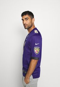 Fialová futbalová dres s čiernymi akcentmi, so zosilneným číslom 8 a logom Baltimore Ravens na rukáve. Vyrobený z priedušnej tkaniny, s krátkymi rukávmi.