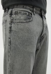 Gros plan sur une personne portant un jean en denim gris délavé avec des poches avant et arrière visibles sur un fond neutre.