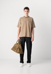 Beige short-sleeve t-shirt with a crew neckline, paired with dark denim trousers. The model holds a brown leather handbag with three front pockets.