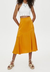 Una falda midi amarilla, fluida, hecha de un tejido suave, presenta un diseño asimétrico con una abertura lateral. Se combina con una blusa de crochet blanca.