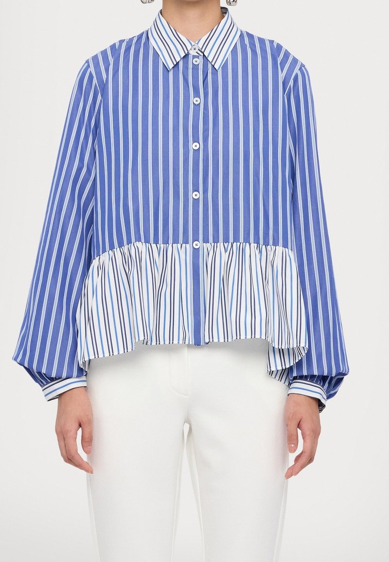 Femme portant un chemisier bleu et blanc rayé verticalement, boutonné, avec un ourlet à volants et un pantalon blanc.