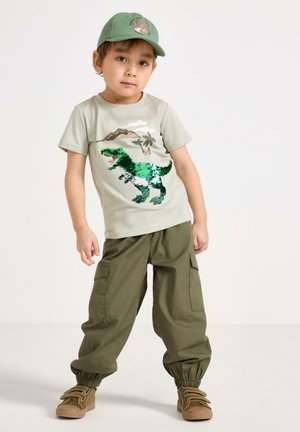 Giovane ragazzo con cappellino verde, t-shirt beige con dinosauro e paillettes, pantaloni cargo oliva e scarpe marroni, in piedi su sfondo bianco.