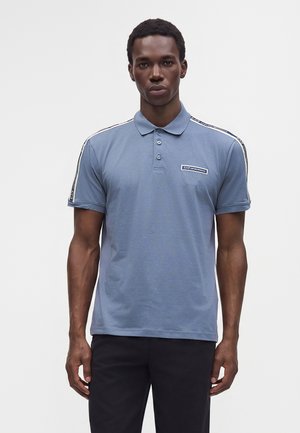 Jeune homme portant un polo bleu avec des bandes contrastantes aux épaules et un écusson logo, associé à un pantalon noir, debout face à l'avant.