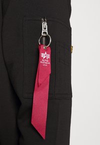 Tela negra con costuras visibles presenta un bolsillo con cremallera y una etiqueta de rayas rojas con la marca "ALPHA INDUSTRIES INC USA".