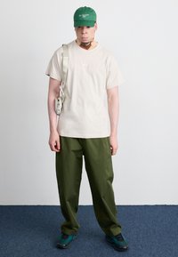 Μπεζ βαμβακερό t-shirt με μικρό λογότυπο, ελαιόχρυσα φαρδιά παντελόνια, πράσινο καπέλο με κεντημένο κείμενο και σχεδίαση αθλητικών παπουτσιών σε τιρκουάζ.