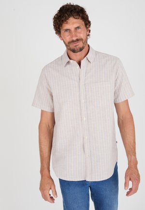 Hombre con cabello rizado y barba que lleva una camisa beige de manga corta con finas rayas blancas y vaqueros azules sobre un fondo liso.